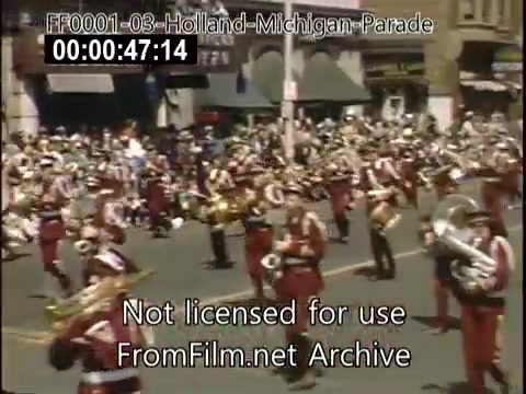 The still captures a vibrant moment from the Holland, Michigan Tulip Parade, circa 1948-1950. A marching band, dressed in red and white uniforms, plays instruments as they parade down a bustling street. Spectators gather along the sidewalks, enjoying the lively atmosphere of the event. The background features storefronts, emphasizing the town's celebration of its Dutch heritage through music and community participation.