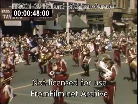 The still depicts a vibrant scene from the Holland, Michigan Tulip Parade, likely between 1948 and 1950. It features a marching band in colorful uniforms performing in front of a large crowd along the street. The atmosphere is festive, highlighting the community's celebration with a backdrop of historical buildings and spectators enjoying the event.