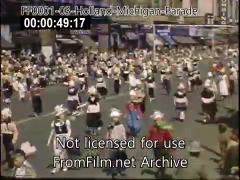 The still captures a vibrant scene from the Holland, Michigan Tulip Parade, likely dating between 1948 and 1950. The image depicts a lively procession filled with participants dressed in traditional attire, contributing to the festive atmosphere. The streets are crowded with spectators enjoying the event. Colorful costumes and hats add to the scene's charm, showcasing the cultural celebration of tulips in the area.