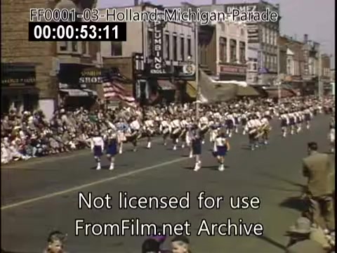 The still captures a lively scene from the Holland, Michigan Tulip Parade, likely between 1948 and 1950. In the foreground, a marching band in uniforms moves along a street filled with spectators. The crowd, ranging from adults to children, watches from the sidewalks amidst storefronts adorned with American flags. The vibrant colors typical of Kodachrome film add to the festive atmosphere of the event.