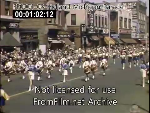 The still captures a lively scene from the Holland, Michigan Tulip Parade, likely between 1948 and 1950. A marching band in coordinated uniforms is parading down the street, accompanied by a large crowd of spectators. Historic storefronts line the background, emphasizing the small-town charm of Holland during this festive event. The vibrant colors typical of Kodachrome film enhance the celebratory atmosphere.