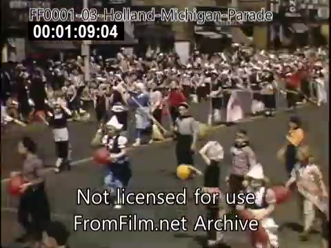The still from 'Holland, Michigan Tulip Parade' captures a lively scene from the late 1940s to early 1950s. The image shows a parade with participants dressed in colorful costumes, playing instruments, and carrying objects such as pom-poms and balls. A large crowd can be seen in the background, highlighting the festive atmosphere of the Tulip Festival, which celebrates Dutch heritage and culture. The vivid colors typical of Kodachrome film convey the vibrancy of the event.