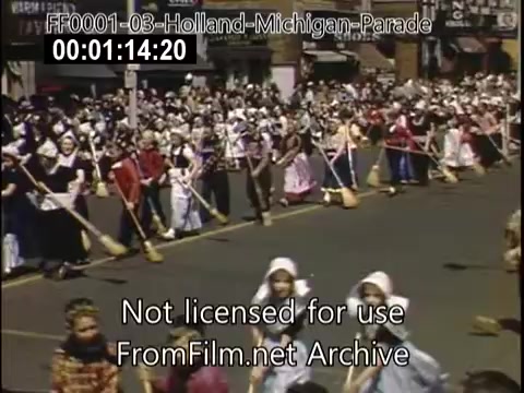 The still captures a vibrant scene from the Holland, Michigan Tulip Parade, circa 1948-1950. Participants, dressed in traditional Dutch costumes, march down a bustling street, showcasing colorful attire. Many hold wooden shoes or brooms, contributing to the festive atmosphere. A large crowd observes, creating a lively and cultural celebration. The image reflects the community's rich heritage and the joy of the annual tulip festival.
