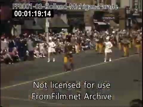 The still features a vibrant scene from the Holland, Michigan Tulip Parade, likely captured between 1948 and 1950. In the foreground, a group of performers in bright costumes leads the procession, while a diverse audience watches from the sidelines. The atmosphere is festive, showcasing the community's celebration with colorful clothing and decorations amidst a backdrop of historical storefronts.