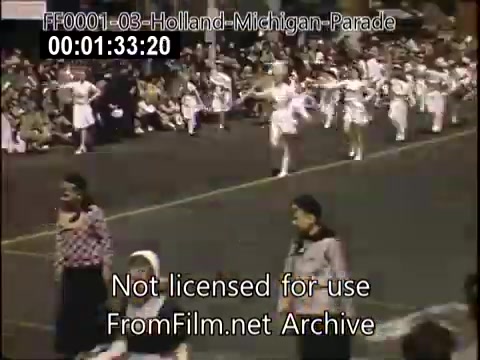 The still captures a moment from the Holland, Michigan Tulip Parade, circa 1948-1950. Featured are participants in colorful costumes, likely including dancers and performers, parading down the street in front of a gathered crowd. The festive atmosphere is enhanced by the vibrant colors typical of Kodachrome footage. The scene reflects the cultural celebration honoring Dutch heritage through traditional attire and activities.