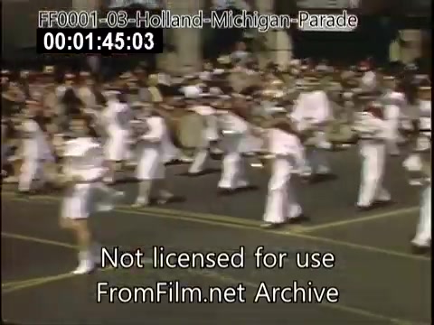 The still depicts a scene from the Holland, Michigan Tulip Parade, filmed in the late 1940s to early 1950s. Participants dressed in white uniforms are marching in formation, showcasing a celebratory atmosphere. A crowd watches from the sidelines, contributing to the festive setting typical of parades. The image captures a nostalgic moment of community celebration.