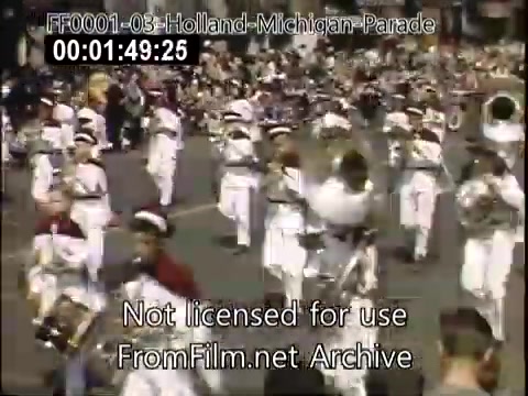 The still captures a lively scene from the Holland, Michigan Tulip Parade, likely between 1948 and 1950. Marching bands dressed in white uniforms perform in formation, while a sizable crowd gathers to watch the festivities. The vibrant Kodachrome colors depict a celebratory atmosphere, highlighting the community's traditions and the picturesque charm of the event.