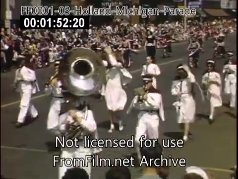 The still captures a lively scene from the Holland, Michigan Tulip Parade, likely between 1948 and 1950. A marching band is prominently featured, with musicians in white uniforms playing brass instruments. The street is filled with spectators, reflecting the festive atmosphere of the parade. The vibrant colors typical of Kodachrome film enhance the sense of celebration inherent in this event.