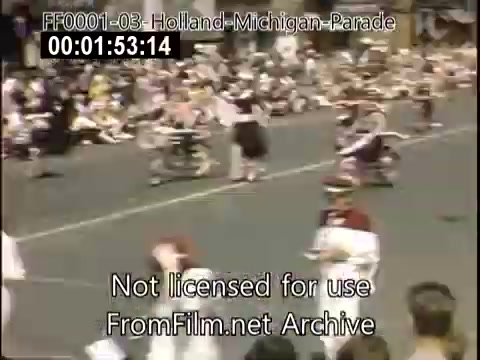 The still captures a vibrant scene from the Holland, Michigan Tulip Parade, circa 1948-1950. It features participants, likely in traditional attire, marching down a lively street. The crowd, made up of spectators, watches from the sidelines, enjoying the festive atmosphere. The Kodachrome film enhances the colors, showcasing the lively energy of the parade.
