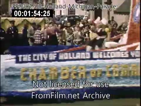 The still captures a vibrant scene from the Holland, Michigan Tulip Parade, circa 1948-1950. It features a colorful float adorned with flowers and a large banner that reads, 'The City of Holland Welcomes You.' Participants in traditional attire can be seen, contributing to the festive atmosphere. The background includes spectators and vintage cars, reflecting the mid-20th century setting.