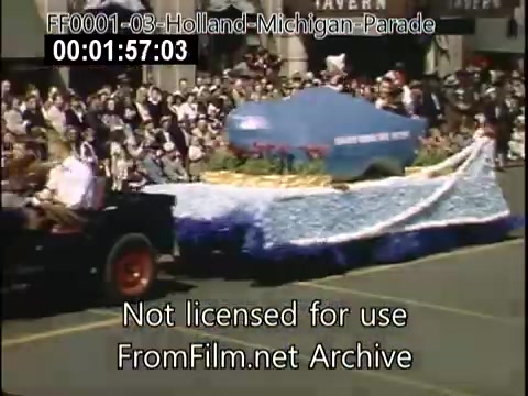 The still captures a scene from the Holland, Michigan Tulip Parade circa 1948-1950. It features a decorated float, possibly representing a car, adorned with vibrant floral arrangements. A crowd watches enthusiastically from the sidelines, and the setting is festive with a clear sunny day. The lively atmosphere is enhanced by the historical context of the early post-war era.