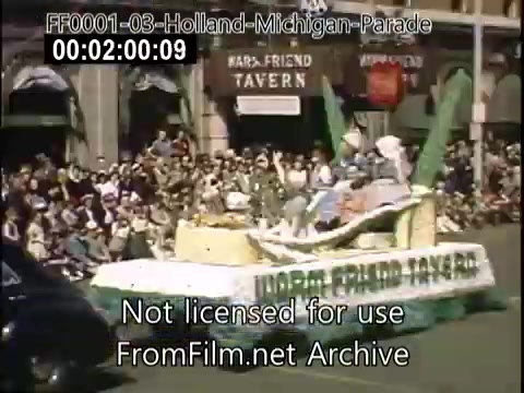 The still captures a vibrant scene from the Holland, Michigan Tulip Parade, dated circa 1948-1950. A float adorned with tulip-themed decorations is featured prominently, showcasing participants dressed in traditional attire. A large crowd of spectators watches from the sidelines, gathered in front of a building marked 'Friend Tavern.' The atmosphere is festive, reflecting the celebration of Dutch heritage and community spirit typical of the event.