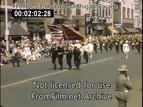 The still captures a moment from the Holland, Michigan Tulip Parade, circa 1948-1950. In the foreground, a group of marchers, possibly including flag bearers, leads the procession along a street lined with spectators. The crowd appears enthusiastic, with a mix of adults and children watching the event. The vintage Kodachrome footage showcases the vibrant colors and festive atmosphere typical of a community celebration featuring floral motifs.
