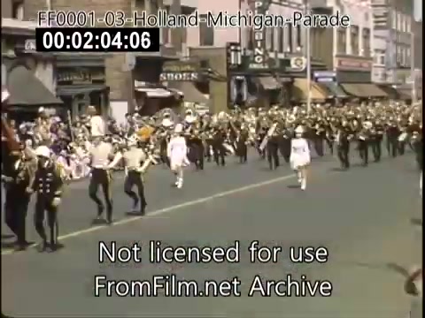The still captures a scene from the Holland, Michigan Tulip Parade, circa 1948-1950. It shows a lively street populated with marching bands and performers in distinctive uniforms. The parade features participants in white outfits, likely representing local schools or organizations, marching down a bustling street lined with spectators and shops. The vibrant colors of the Kodachrome footage enhance the festive atmosphere of the event.