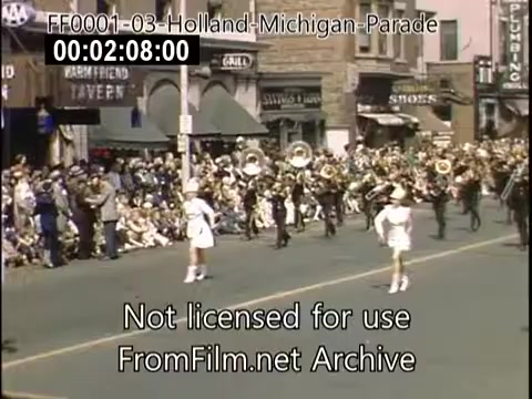 The still from the 'Holland, Michigan Tulip Parade' (circa 1948-1950) captures a vibrant scene featuring a marching band on a lively street. Spectators line the sides, eagerly watching the procession, while a young female performer in a white outfit leads the way. The backdrop includes vintage storefronts and colorful signs, typical of mid-20th century America, enhancing the festive atmosphere of the parade.