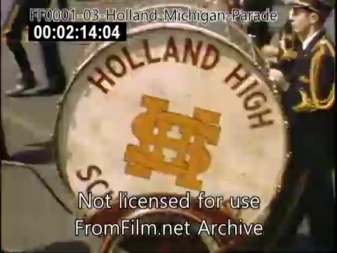 The still depicts a vibrant scene from the Holland, Michigan Tulip Parade, likely taken between 1948 and 1950. A prominent feature is the large drum of the Holland High School marching band, showcasing the school's name in bold letters. The drum is decorated with a bright orange and yellow design, adding to the festive atmosphere of the parade. The background hints at a lively crowd and other parade participants, characteristic of local celebrations celebrating Dutch heritage.