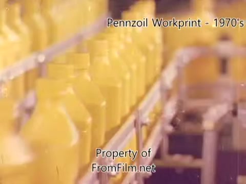 The still from the 'Pennzoil Factory/Refinery Workprint - 1980s' shows a close-up view of yellow bottles lined up on a production line. The film exhibits a vintage aesthetic typical of 16mm negatives, with a slight grain and some debris visible in the film gate. The bottles appear uniform and are likely indicative of the oil or automotive fluid being produced. The background has a blurred effect, emphasizing the bottles in the foreground, conveying a sense of industrial activity. The accompanying music from Underbelly enhances the nostalgic atmosphere of the footage.