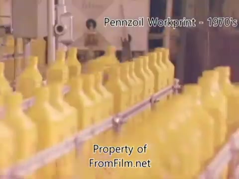 The still from the 'Pennzoil Factory/Refinery Workprint - 1980s' shows a manufacturing scene with numerous yellow oil bottles on a production line. The image captures a moment during the bottling process, highlighting the factory's industrial environment. Some debris is visible in the film gate, indicative of the age and condition of the 16mm negative. The music accompanying the film is from Underbelly and includes tracks like 'I Hear Voices,' 'Asterism,' 'Italy Jr.,' and 'Land's End.'