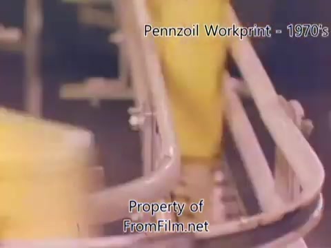 The still from the 'Pennzoil Factory/Refinery Workprint - 1980s' showcases a segment of the manufacturing process, with a focus on machinery and conveyor belts in action. The film exhibits some noticeable debris in the gate, which is common in older 16mm negatives. Accompanying this visual, the music from Underbelly adds an atmospheric layer, featuring tracks like 'I Hear Voices,' 'Asterism,' 'Italy Jr.,' and 'Land's End.' The overall aesthetic reflects the industrial setting of the Pennzoil refinery.