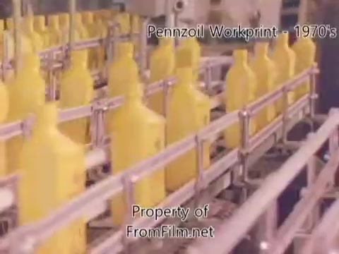 The still shows a production line at the Pennzoil refinery facility, featuring numerous yellow bottles lined up on a conveyor belt. The setting suggests an industrial environment focused on packaging motor oil. The film is a workprint from the 1980s, with visible debris in the film gate indicating age. Accompanying music includes tracks from Underbelly, adding a contemporary feel to the retro imagery.
