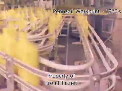 The 16mm still from the 'Pennzoil Factory/Refinery Workprint' depicts a scene from the 1970s showing yellow bottles moving along a conveyor system in a refinery setting. The image captures the industrial environment typically associated with oil production, highlighting the machinery and processes involved. The film has noticeable debris in the gate, indicating its age. The backdrop features metallic structures characteristic of refinery architecture. The accompanying music by Underbelly provides an atmospheric layer to the visual elements.
