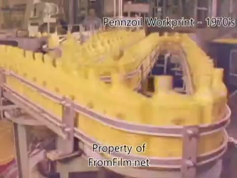 The still shows a production line in a Pennzoil refinery during the 1980s. Yellow oil bottles are prominently featured on a conveyor belt, indicating the bottling process. The film exhibits some debris in the film gate, typical of older film stock. The background suggests an industrial setting, emphasizing the manufacturing aspect of the refinery. The accompanying music from Underbelly adds an energetic atmosphere to the workprint.