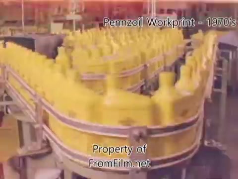 The still shows a production line in a Pennzoil refinery from the 1970s, featuring yellow bottles of oil in a circular arrangement on a conveyor belt. The image captures the industrial setting, highlighting the manufacturing process. Debris is noted in the film gate, providing a vintage feel typical of 16mm workprints. The accompanying music features tracks from Underbelly, enhancing the nostalgic atmosphere.