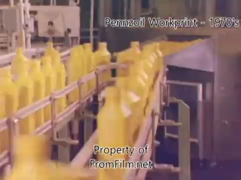 The still captures a scene from a 16mm workprint of a Pennzoil refinery from the 1970s. It features a production line with yellow oil containers moving along a conveyor belt. Background elements suggest a factory setting, typical of oil processing. The film has noticeable debris in the film gate, indicating its age and use. The accompanying music by Underbelly adds an ambient atmosphere to the industrial visuals.