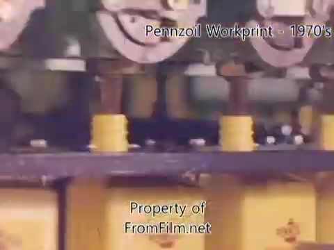 The still from the 'Pennzoil Factory⧸Refinery Workprint' showcases machinery involved in the oil production process. It captures a close-up view of equipment with yellow oil containers, likely highlighting the bottling or processing stage. The image appears to have some debris in the film gate, indicating it's a workprint from a 16mm negative, typical of the 1970s aesthetic. The accompanying music by Underbelly contributes to the overall ambiance of the footage.