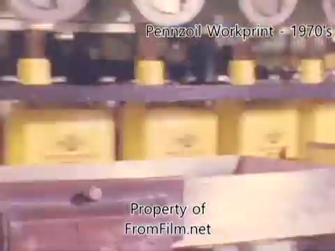 The still from 'Pennzoil Factory/Refinery Workprint - 1980's' features a close-up view of yellow Pennzoil containers on a production line, highlighting the factory setting. The film has visible debris in the gate, characteristic of 16mm negatives, emphasizing its vintage nature. The atmosphere captures the essence of industrial production, complemented by music from Underbelly, enhancing the nostalgic feel of the workprint.