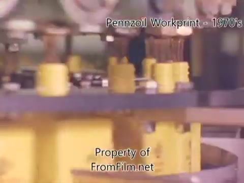 The still shows a close-up view of machinery within a Pennzoil refinery or factory, specifically focusing on yellow containers or components being processed. The film exhibits some debris in the film gate, emphasizing the wear typical of a 16mm workprint. The context is rooted in industrial work from the 1980s, accompanied by music tracks from Underbelly, contributing to an evocative atmosphere of the era.