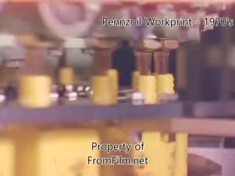 The still from 'Pennzoil Factory/Refinery Workprint - 1980's' captures a close-up view of industrial machinery within a refinery setting. The image features yellow components, likely valves or fittings, against a backdrop that suggests a complex assembly or processing environment. Debris is visible in the film gate, indicating aging or wear in the film. The accompanying music by Underbelly provides an atmospheric backdrop to the visual narrative of industrial activity from the era.