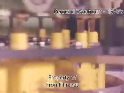 The still from the 'Pennzoil Factory/Refinery Workprint - 1980s' presents a close-up view of industrial machinery, featuring yellow components prominently. The image suggests a focus on the operational aspects of the refinery, typical of the period's industrial documentation. Debris can be seen in the film gate, indicating age and wear typical of a 16mm negative. The accompanying music by Underbelly enhances the atmosphere, likely providing a backdrop to the visual narrative of the refinery's work processes.