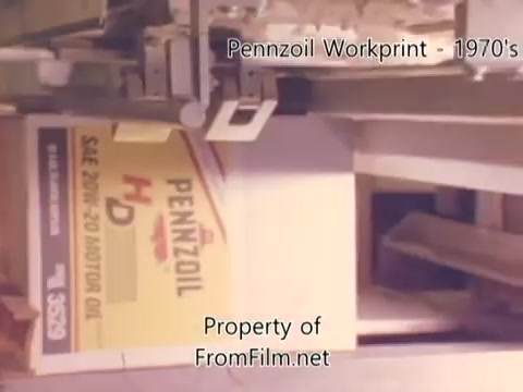 The still depicts machinery in a refinery setting, focused on a box labeled 'Pennzoil HD SAE 20W-30 MOTOR OIL.' The image captures a moment during the manufacturing process, with visible debris in the film gate, likely due to age. The overall aesthetic reflects a 1980s workprint style, with a somewhat muted color palette common in older film formats. The accompanying music tracks by Underbelly add a distinct audio backdrop to the visuals.