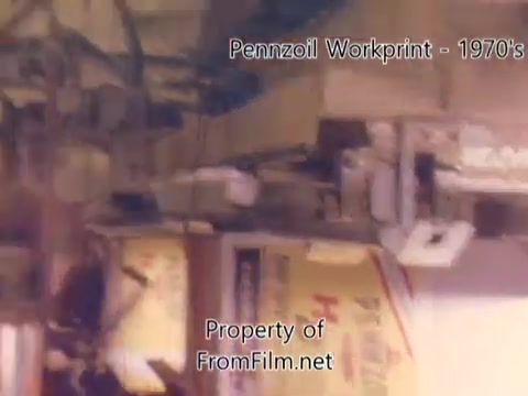 The still from the 'Pennzoil Factory / Refinery Workprint - 1980's' showcases a scene from the manufacturing process, likely within a refinery setting. It features machinery, equipment, and packaging elements typical of industrial environments. Some debris is visible in the film gate, indicating wear or age in the film rather than the projector. The accompanying music enhances the nostalgic atmosphere, with tracks from Underbelly adding a distinct auditory layer to the visual experience.