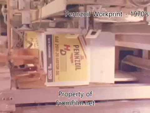 The 16mm still from 'Pennzoil Factory⧸Refinery Workprint - 1980's' depicts machinery involved in the production or packaging of Pennzoil products. The image shows boxes labeled with the Pennzoil branding, indicating a focus on oil or lubricant production. Notable debris in the film gate suggests an aged film print, typical of workprints from that era. Accompanying music from Underbelly enhances the industrial ambiance of the scene.