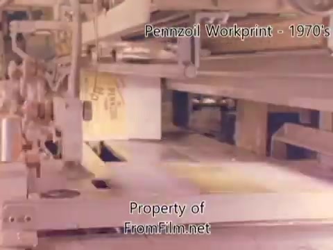 The still from the 'Pennzoil Factory/Refinery Workprint - 1980s' features industrial machinery at a Pennzoil facility. The scene captures a segment of the production process, showcasing the inner workings of the refinery. The equipment appears to be engaged in processing or packaging oil products. Notably, there is visible debris in the film gate, indicating wear on the 16mm film negative. The accompanying music by Underbelly includes tracks like 'I Hear Voices,' enhancing the ambiance of the footage.