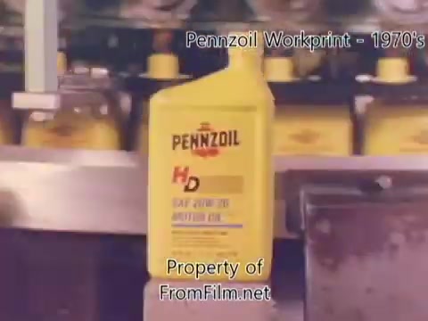 The still depicts a scene from a Pennzoil factory or refinery workprint from the 1980s, showcasing a container of Pennzoil HD motor oil prominently in the foreground. The background features rows of yellow Pennzoil packaging, emphasizing the brand's production environment. The image has some visible debris in the film gate, indicative of the film's age and condition. The scene is likely accompanied by music from Underbelly, adding to the nostalgic feel of the workprint.