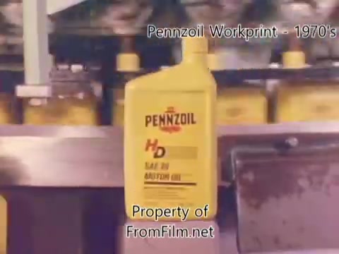 The still features a prominent yellow Pennzoil bottle labeled 'HD' against a backdrop of a refinery or factory setting. The packaging reflects the branding and design typical of the 1970s. Debris is visible in the film gate, indicating wear typical of a workprint. The visual captures the industrial atmosphere, likely showcasing the bottling process, enhancing the context of quality oil production.