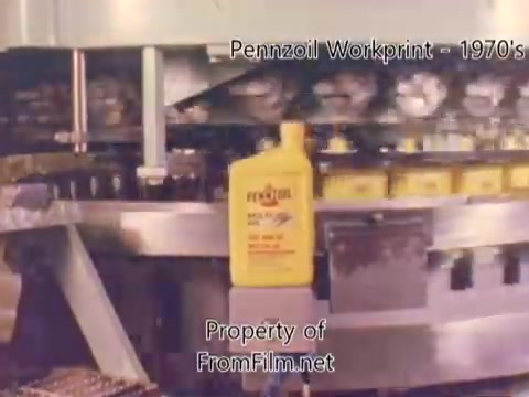 The still from 'Pennzoil Factory/Refinery Workprint - 1980's' depicts a production line showcasing yellow Pennzoil bottles being processed. The focus is on a singular yellow bottle prominently placed in the frame, likely highlighting the brand. The background features industrial machinery and other bottles moving along a conveyor system, illustrating the manufacturing process. Debris can be seen in the film gate, indicating age or wear of the film. The accompanying music by Underbelly adds a modern auditory layer to the vintage visuals.