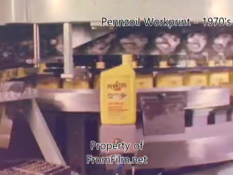 The still depicts a segment from a 16mm workprint of the Pennzoil refinery, likely from the 1970s. In the image, containers of Pennzoil are visible on a moving conveyor system, highlighting the industrial setting of the factory. Some debris can be seen in the film gate, which is part of the film's condition rather than the projection equipment. The accompanying music is by Underbelly, featuring tracks that complement the visual elements of the refinery work.