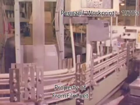 The 16mm still from the 'Pennzoil Factory/Refinery Workprint - 1980's' depicts the interior of a Pennzoil refinery. The image shows industrial machinery and equipment, likely part of the oil production process. The film has visible debris in the gate, which is noted not to be a projector issue. Accompanying music from Underbelly enhances the industrial and historical atmosphere.