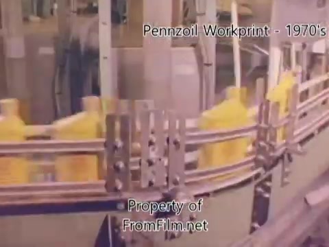 The still from the 'Pennzoil Factory/Refinery Workprint - 1980's' depicts a conveyor belt transporting yellow oil containers, characteristic of a manufacturing or refining process. The film exhibits some debris in the gate, a common occurrence in vintage film. The context suggests an insight into industrial operations, enhanced by a soundtrack from the YouTube Audio Library featuring tracks like 'I Hear Voices' and 'Land's End.'