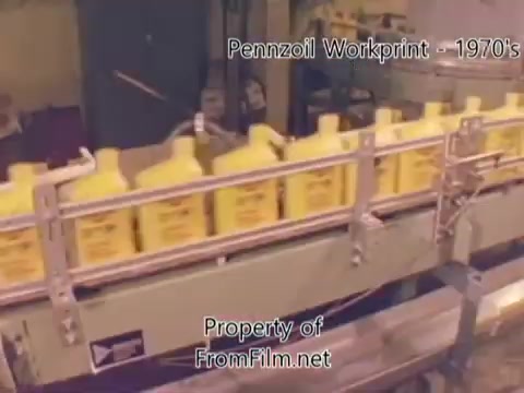 The still depicts a production line from the Pennzoil factory, featuring yellow oil containers moving along a conveyor belt. The environment appears industrial, typical of a refinery setting. Notable are the visible debris in the film gate, suggesting wear on the 16mm negative. The music overlay includes tracks from Underbelly, enhancing the nostalgic feel of the workprint.