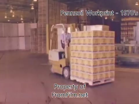 The still from the 'Pennzoil Factory⧸Refinery Workprint - 1980's' features a forklift transporting a pallet stacked with boxes, likely containing oil products. The setting appears to be an industrial environment, typical of a refinery or factory, with visible stacks of materials in the background. The film grain and some debris in the film gate contribute to its vintage aesthetic. Accompanying music from Underbelly enhances the nostalgic feel of this workprint.