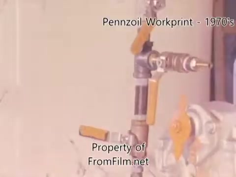 The still shows a close-up of industrial machinery from a Pennzoil refinery, likely from the 1980s. The equipment features metallic pipes and valves, with some yellow handles visible. There is noticeable debris in the film gate, which affects the image quality. Accompanying music is by Underbelly, featuring tracks such as 'I Hear Voices' and 'Asterism.' The workprint captures a raw, authentic glimpse into the refinery's operations.