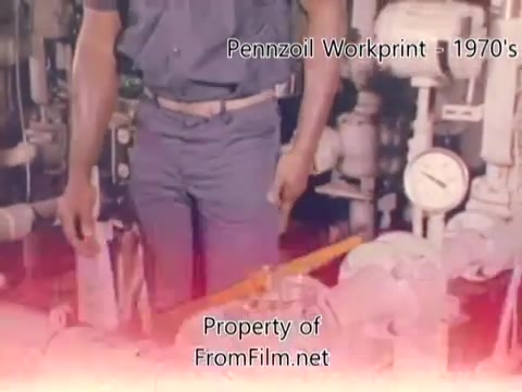 The still from the 'Pennzoil Factory/Refinery Workprint - 1980's' shows a worker standing in front of machinery typical of an industrial setting, likely within a refinery. The focus appears to be on the individual, whose arms are positioned near mechanical gauges and controls, indicating their role in operations or monitoring. The film has a vintage look characteristic of 16mm negatives, with some visible debris in the film gate. The background is blurred with a subtle color gradient, emphasizing the worker. The music accompanying this footage includes various tracks from Underbelly, enhancing the retro ambiance of the scene.