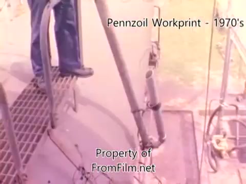 The still depicts a scene from the Pennzoil refinery workprint from the 1980s, showing a worker standing on a metal grate, likely near machinery or equipment. The film shows signs of wear, with noticeable debris in the film gate. The image captures a moment in an industrial environment, emphasizing the manufacturing process associated with refining oil. The soundtrack features tracks by Underbelly, creating an atmospheric backdrop for the visual content.