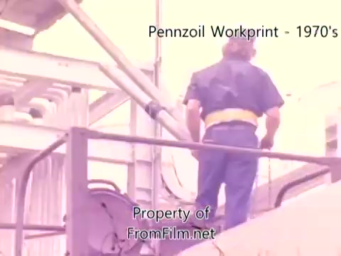 The still from 'Pennzoil Factory/Refinery Workprint - 1980's' features a worker in a blue uniform and yellow belt, positioned on a platform amidst industrial structures. The setting appears to be a refinery, with pipes and machinery visible in the background, showcasing a working environment. The image is part of a 16mm negative workprint, indicating it may not have the clarity of a final cut. Debris can be seen in the film gate, affecting the visual quality. The accompanying music by Underbelly adds an atmospheric backdrop to the industrial scene.