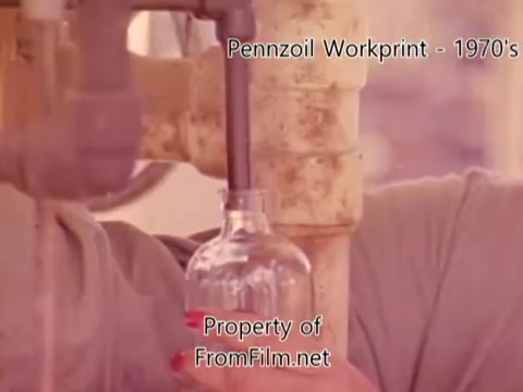 The still from the 'Pennzoil Factory⧸Refinery Workprint - 1980's' depicts a close-up of a person in a factory setting, engaging with machinery while filling a glass bottle. The colors have a warm tint, typical of older film stock, with some visible debris in the film gate, which suggests age and wear. The context indicates it’s a workprint, showcasing industrial processes, and it features music from Underbelly's tracks, enhancing the atmosphere of the scene.
